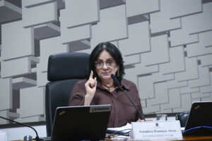 Senadora Damares Alves aparece com camisa cor marrom, com dedo em riste, sentada na mesa da presidência da Comissão de Assuntos Econômicos. Ao fundo, um painel com blocos sobrepostos.